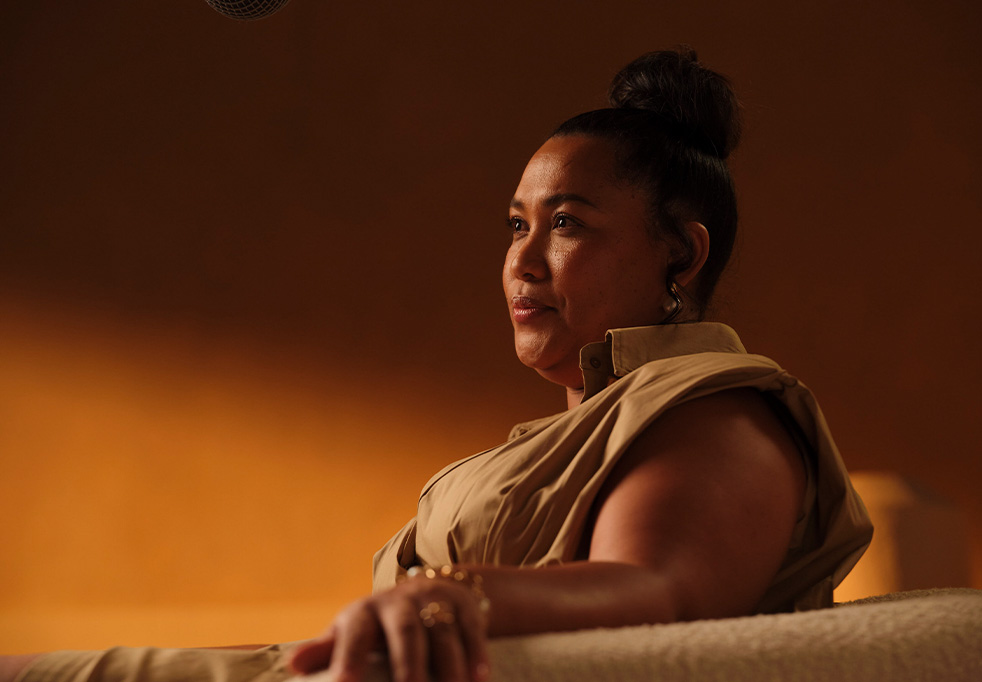 Natasha, a woman with dark hair in a bun, wearing a sleeveless beige dress, sitting in a chair and looking slightly to the side with a calm expression